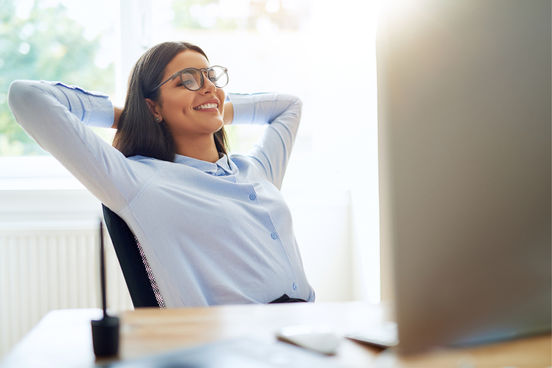 Smiling woman wearing a blue smart shirt sits back relaxed on her desk, with her hands behind her back