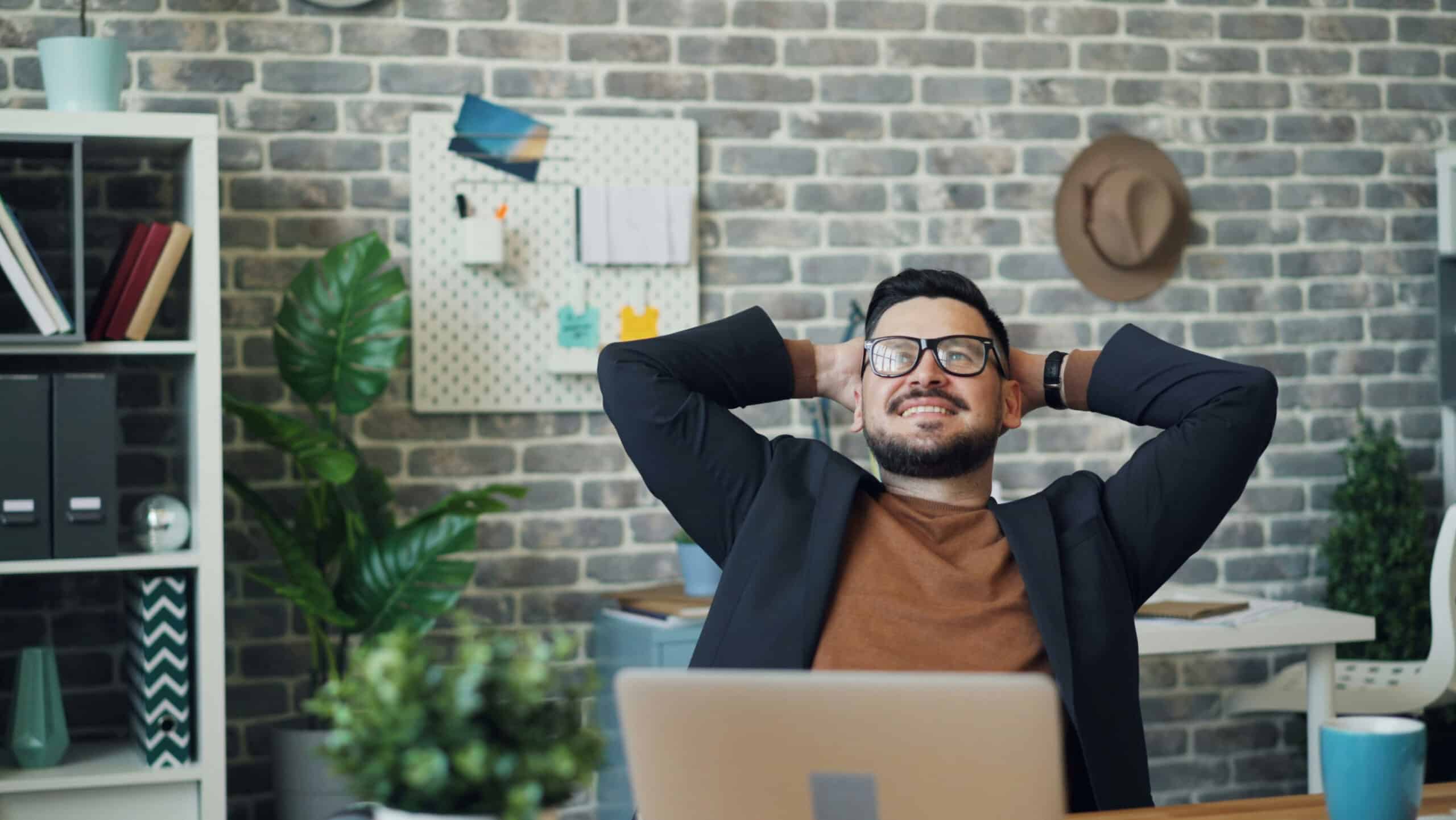 A happy man in an office setting leans back in his chair with his hands behind his back, sat in front of his laptop.